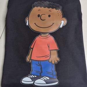 May include: Black t-shirt featuring a cartoon illustration of a smiling boy wearing earbuds. The boy is depicted in a red shirt, blue jeans, and black and white sneakers. The graphic is centered on the shirt.