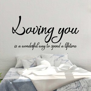 May include: A bedroom scene with a white wall featuring a black vinyl decal that reads "Loving you is a wonderful way to spend a lifetime." The bed has white and blue bedding, with a green plant in a pot on the left.