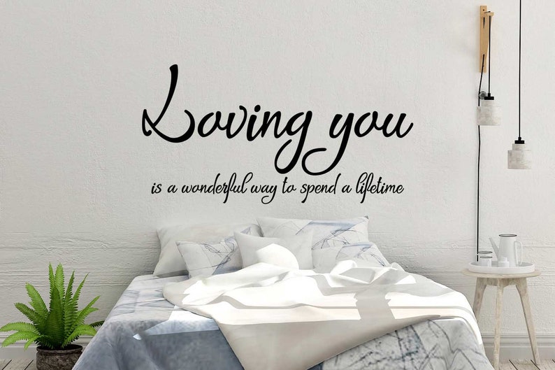 May include: A bedroom scene with a white wall featuring a black vinyl decal that reads "Loving you is a wonderful way to spend a lifetime." The bed has white and blue bedding, with a green plant in a pot on the left.