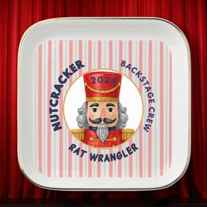 May include: A square white tray with a gold rim featuring a pink and white vertical striped background. In the center, a circular graphic shows a nutcracker wearing a red hat labeled 2026, surrounded by the text Nutcracker Backstage Crew Rat Wrangler.