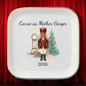 May include: A square white ceramic tray with a gold rim featuring a watercolor illustration of a nutcracker, a grandfather clock, a mouse, and a Christmas tree. The text reads Carrie as Mother Ginger and the year 2026 in black script and font.