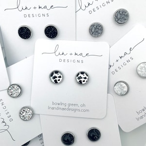May include: Stud earrings in various designs, including black glitter, cow print, and marble patterns. Each pair is mounted on a white card with the "lin + mae DESIGNS" logo and website address. The earrings have a silver-colored metal border.