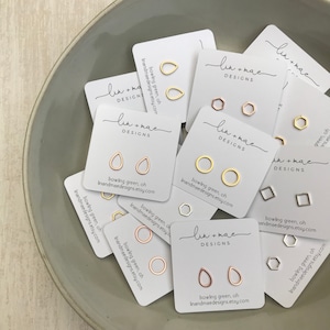 May include: Assortment of small gold and silver earrings on white cards with the text "lin + mae DESIGNS" and "bowling green, oh linandmaedesigns.etsy.com".