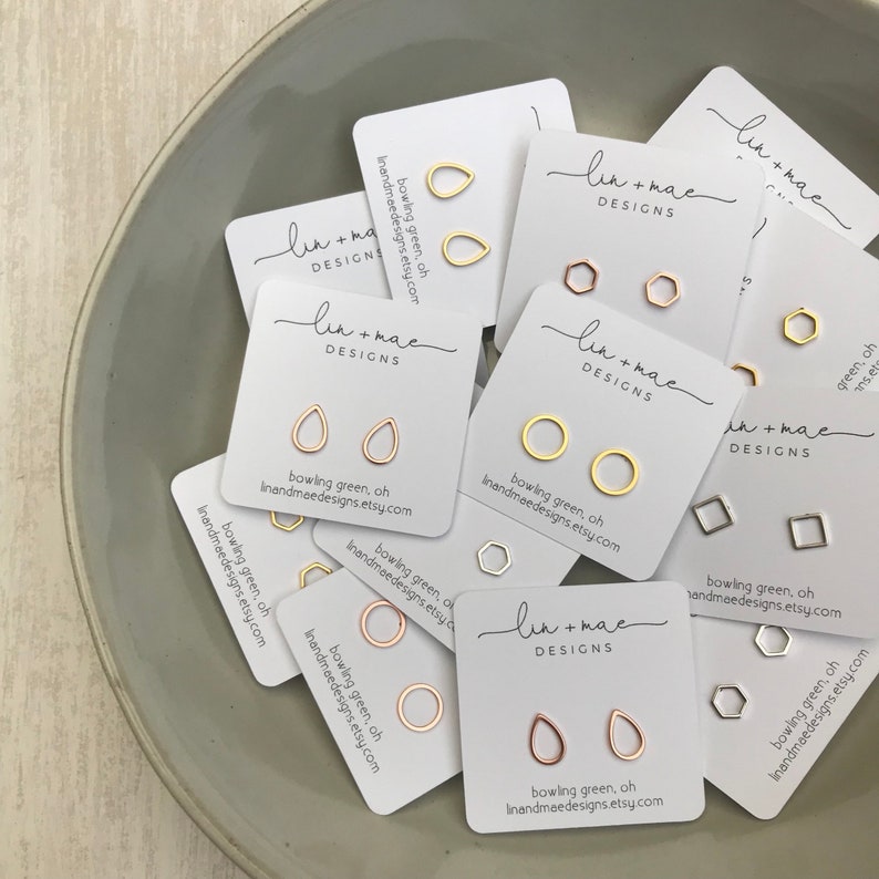May include: Assortment of small gold and silver earrings on white cards with the text "lin + mae DESIGNS" and "bowling green, oh linandmaedesigns.etsy.com".