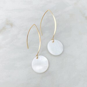 May include: A pair of gold-colored drop earrings with a curved hook design. Each earring features a small, round, white, iridescent pendant. The earrings are displayed on a white and gray marble surface.