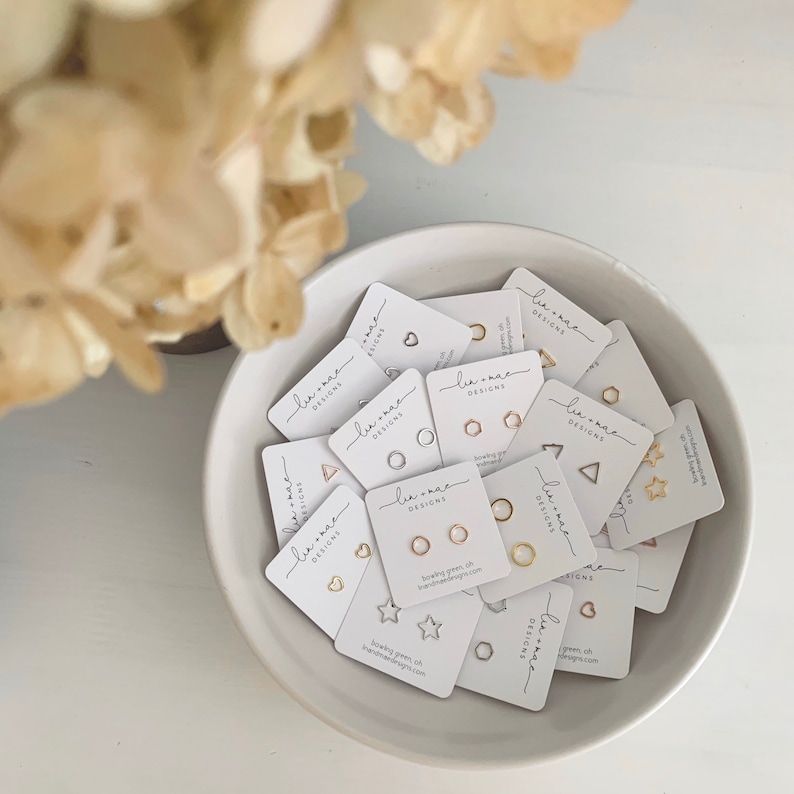 May include: A collection of small, white cards with the text "Le-Mac Designs" printed on them. Each card features a different geometric shape cutout, including a heart, circle, star, triangle, and hexagon. The cards are arranged in a white bowl.