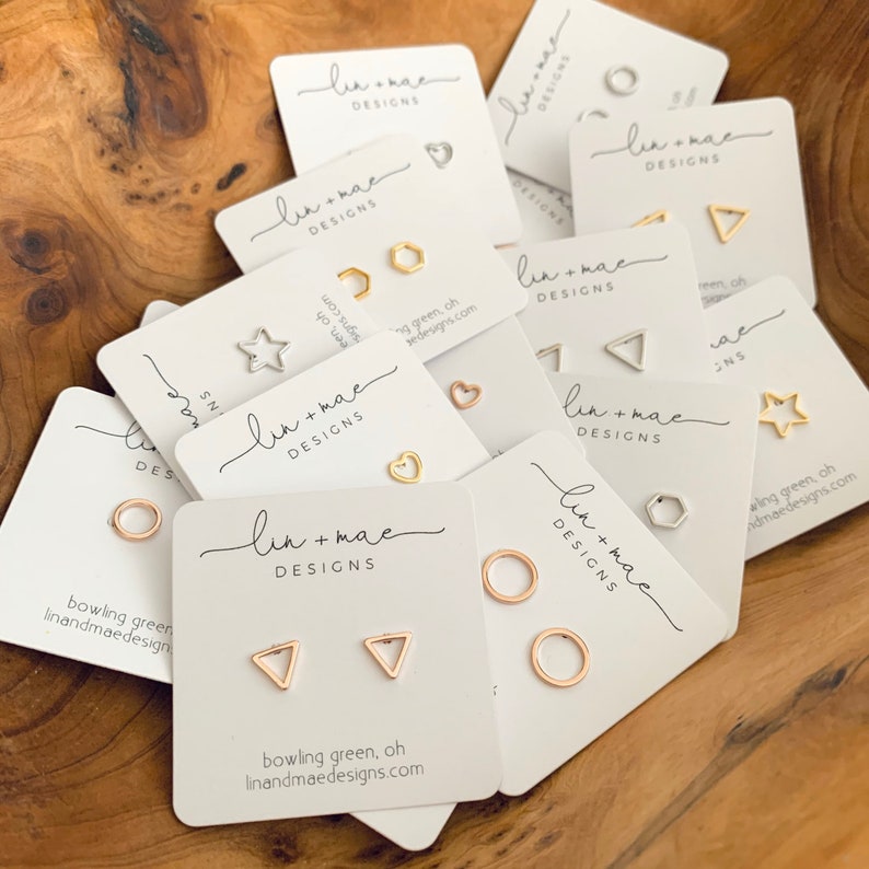 May include: A collection of small, white cards with the text "lin+mae DESIGNS" printed on them. Each card features a pair of gold or silver earrings with geometric shapes, including circles, triangles, stars, and hexagons. The cards are arranged on a wooden surface.