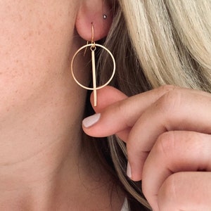 May include: Gold hoop earrings with a vertical bar hanging from the hoop.