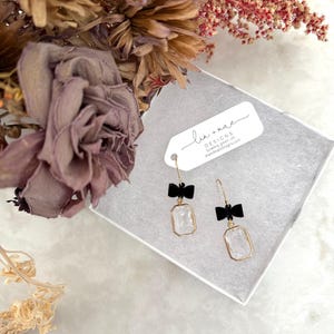 May include: A pair of gold earrings with clear crystal stones and black bows. The earrings are displayed in a white box with a white tag that reads "lin + mae DESIGNS bowing green with intention linandmaedesigns.com".