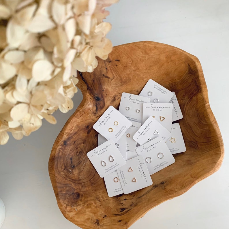 May include: A wooden bowl filled with small cards featuring various geometric shapes. Each card has the text "Lee-Rae Designs" and a small image of a pair of earrings in a different shape. The shapes include a circle, heart, triangle, star, hexagon, teardrop, and a square.