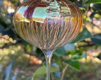 May include: A clear glass ornament with a bulbous, ribbed body and a teardrop-shaped bottom. The ornament has a gold and purple iridescent finish, and is suspended against a backdrop of green foliage.