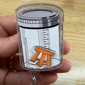 Può includere: Un porta badge in acrilico trasparente con bordo glitterato e una stampa raffigurante un contenitore per campioni con coperchio grigio e tacche di misurazione. Sulla parte anteriore del contenitore è applicata una piccola illustrazione di uno sgabello arancione.
