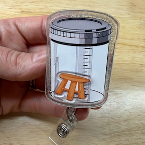 May include: A clear acrylic badge reel with a cartoon image of a medical urine collection container and a small orange stool. The badge reel has a silver-colored metal clip and a clear plastic strap.