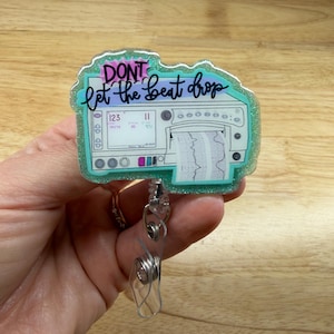 Peut inclure: Un porte-badge scintillant turquoise avec le texte "DON'T let the beat drop" en écriture noire. Le badge présente un motif de moniteur médical avec un tracé de fréquence cardiaque. Un enrouleur en plastique transparent et un clip en métal sont attachés.