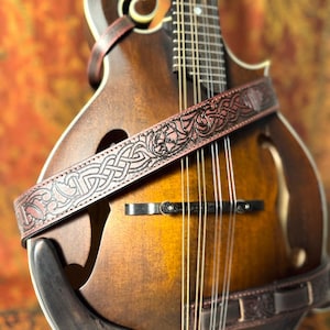 May include: A close-up of a brown mandolin with a decorative leather strap. The strap features an embossed Celtic knot design in a reddish-brown color. The mandolin has a wooden body with a sunburst finish and metal strings.