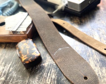 May include: A close-up of a brown leather guitar strap with a textured surface. The strap has a hole at the end and a brand name printed on it. Other guitar accessories are in the background.