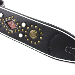 Pu&ograve; includere: Cinturino per chitarra in pelle nera con cuciture bianche e borchie dorate. Il cinturino presenta un design a forma di diamante con un intarsio in pelle marrone e un emblema metallico circolare con un design. Il testo "WILD BILLY'S" &egrave; in rilievo sul cinturino.