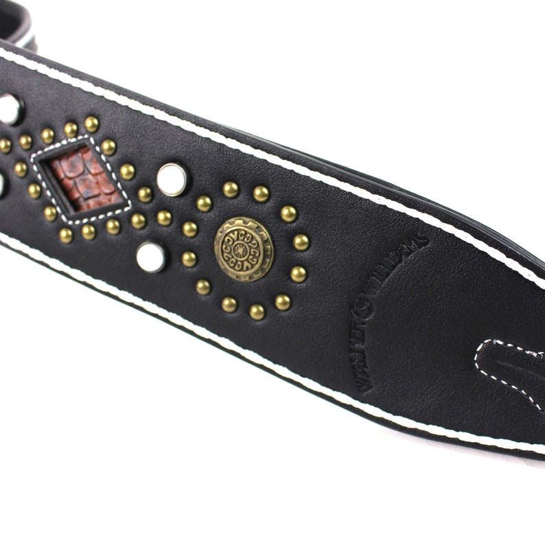 Pu&ograve; includere: Cinturino per chitarra in pelle nera con cuciture bianche e borchie dorate. Il cinturino presenta un design a forma di diamante con un intarsio in pelle marrone e un emblema metallico circolare con un design. Il testo "WILD BILLY'S" &egrave; in rilievo sul cinturino.