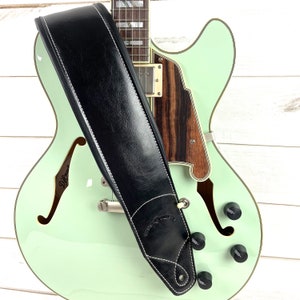 May include: A black leather guitar strap with white stitching is draped over a mint green electric guitar. The strap has a padded design and features the text "Walker & Williams". The guitar has a wooden pickguard and black knobs.