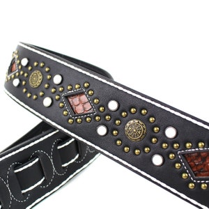 Pu&ograve; includere: Cinturino per chitarra in pelle nera con cuciture bianche e borchie decorative, tra cui accenti metallici rotondi e pezzi a forma di diamante con un motivo in pelle marrone.