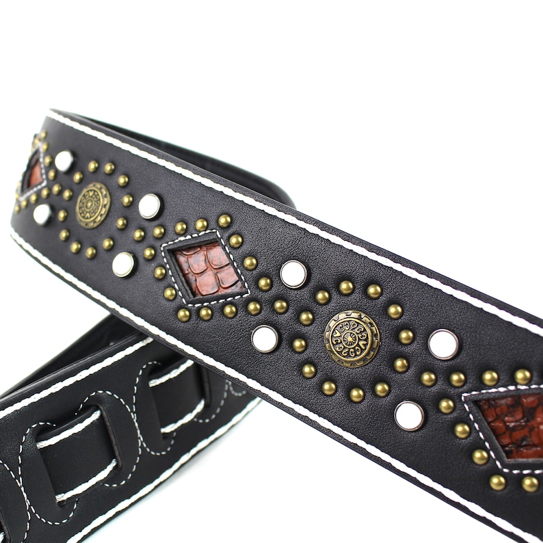 Pu&ograve; includere: Cinturino per chitarra in pelle nera con cuciture bianche e borchie decorative, tra cui accenti metallici rotondi e pezzi a forma di diamante con un motivo in pelle marrone.