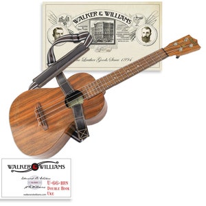 May include: A brown wooden ukulele with a black leather strap. The ukulele has a double hook and is made by Walker & Williams. The image also includes a vintage advertisement for Walker & Williams leather goods.