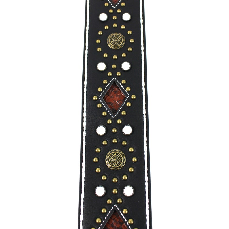 Pu&ograve; includere: Cinturino per chitarra in pelle nera con cuciture bianche e borchie e dettagli dorati. Il cinturino ha un motivo a forma di diamante con un inserto in pelle marrone.