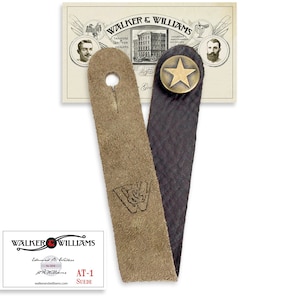 May include: A brown leather guitar strap with a star-shaped metal button and the text "WALKER & WILLIAMS" embossed on the strap. The strap is attached to a card with the text "WALKER & WILLIAMS Established 1894 J.H. Williams walkerandwilliams.com AT-1 SUEDE".