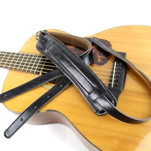 May include: A black leather guitar strap with a padded shoulder section rests on a wooden acoustic guitar. The strap has adjustable length and is designed to attach to the guitar's body. The guitar has a natural wood finish.