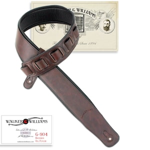 Puede incluir: Correa de guitarra de cuero marr&oacute;n con un patr&oacute;n decorativo y una parte inferior acolchada negra. La correa est&aacute; etiquetada "G-904 Bourbon Oil Finish" y "Walker & Williams Established 1894".