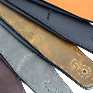 Puede incluir: Primer plano de cuatro correas de guitarra de cuero en diferentes colores: marr&oacute;n, gris, negro y naranja. Las correas tienen un borde cosido y un peque&ntilde;o lazo de metal. Las correas est&aacute;n etiquetadas con "Willie & Willy".