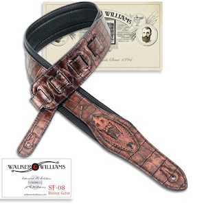 May include: A brown leather guitar strap with a crocodile pattern and an embossed alligator head design. The strap is made by Walker & Williams and is labelled "SF-08 Bronze Gator".