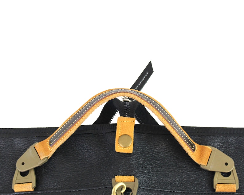 May include: Close-up of a black leather bag with a tan leather handle and a zipper closure. The handle has a brown stitching detail.