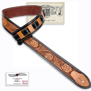 May include: A brown leather guitar strap with black leather accents and floral tooling. The strap has a black leather buckle and a white stitching detail. The strap is made by Walker & Williams.