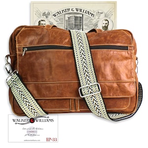 May include: A brown leather messenger bag with a black and white patterned strap. The bag has a zippered pocket on the front and a flap closure. The bag is sitting on a white surface. The bag is labeled "Walker & Williams" and "HP-33".