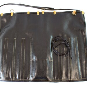 May include: A black leather roll with multiple slots for holding tools or utensils. The roll has a strap with a buckle and gold metal accents.