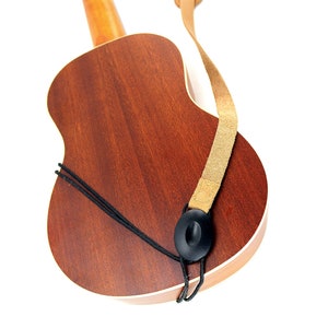 Peut inclure: Un ukul&eacute;l&eacute; brun avec une sangle en cuir beige attach&eacute;e &agrave; l'instrument. La sangle a un bouton noir &agrave; l'extr&eacute;mit&eacute;.