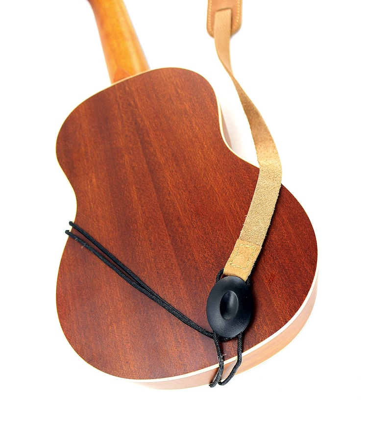 Peut inclure: Un ukul&eacute;l&eacute; brun avec une sangle en cuir beige attach&eacute;e &agrave; l'instrument. La sangle a un bouton noir &agrave; l'extr&eacute;mit&eacute;.
