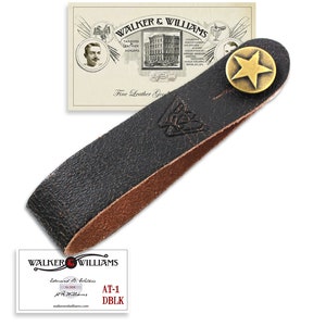 May include: A black leather guitar strap with a brown leather edge and a brass star-shaped button. The strap is embossed with the text "Walker & Williams" and a logo. The strap is labeled "AT-1 DBLK".