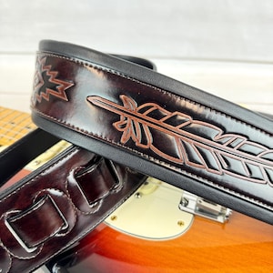 May include: A brown leather guitar strap with a black border and a brown leather feather design. The strap is on top of a guitar.