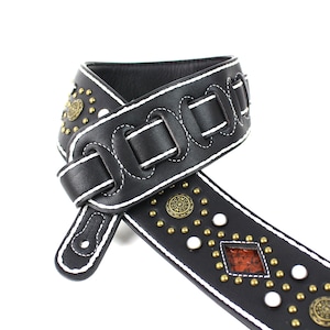Pu&ograve; includere: Cinturino per chitarra in pelle nera con cuciture bianche e dettagli metallici decorativi. Il cinturino ha un design unico con pi&ugrave; file di borchie e un motivo a cerchi.