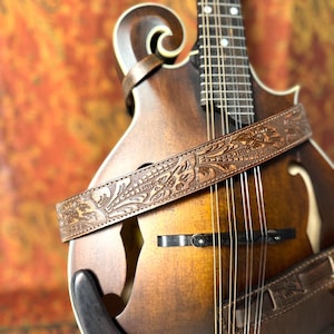 May include: A brown mandolin with a decorative, embossed leather strap. The strap features a detailed floral and geometric pattern. The instrument has a wooden body with a sunburst finish and metal strings. The background is a warm, textured orange.