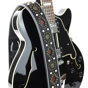 Pu&ograve; includere: Una chitarra elettrica nera con una tracolla in pelle bianca e marrone con borchie decorative e dettagli in metallo.