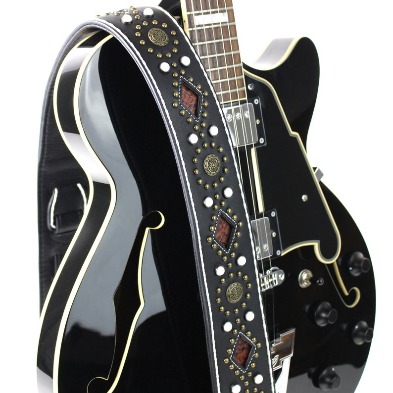 Pu&ograve; includere: Una chitarra elettrica nera con una tracolla in pelle bianca e marrone con borchie decorative e dettagli in metallo.