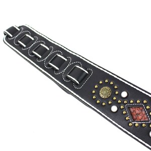 Pu&ograve; includere: Cinturino per chitarra in pelle nera con cuciture bianche e dettagli metallici decorativi. Il cinturino ha un design a forma di diamante con un motivo rosso e marrone.