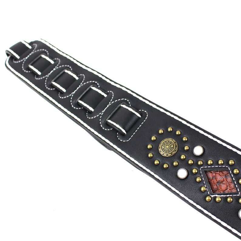 Pu&ograve; includere: Cinturino per chitarra in pelle nera con cuciture bianche e dettagli metallici decorativi. Il cinturino ha un design a forma di diamante con un motivo rosso e marrone.