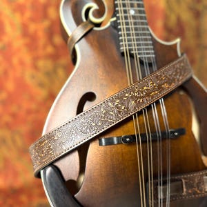 May include: A close-up of a brown mandolin with a decorative, embossed leather strap. The strap features a floral pattern and wraps around the instrument's body. The mandolin has a dark wood finish and visible strings.