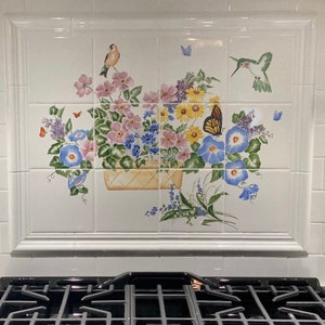May include: A white tile backsplash with a painted floral design. The design features a basket overflowing with pink, blue, yellow, and white flowers. Two hummingbirds are flying above the basket, and butterflies are scattered throughout the design.