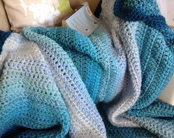 May include: A soft, blue and white crocheted blanket with a gradient of color. The blanket is folded and draped over a couch.