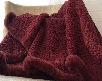 May include: A burgundy throw blanket with a textured, woven design and a plush, faux fur trim. The blanket is draped over a chair, showcasing its soft texture and rich color. A cozy home decor item.